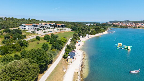 Vue aérienne d’un hôtel situé sur la côte, avec une plage, un port de plaisance et une zone boisée.