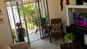 A living room with a sofa, a television, a glass coffee table, and a sliding glass door leading to a garden. - Welcome to Casa Trajano in Olinda! Here, you will find comfort! (Olinda)