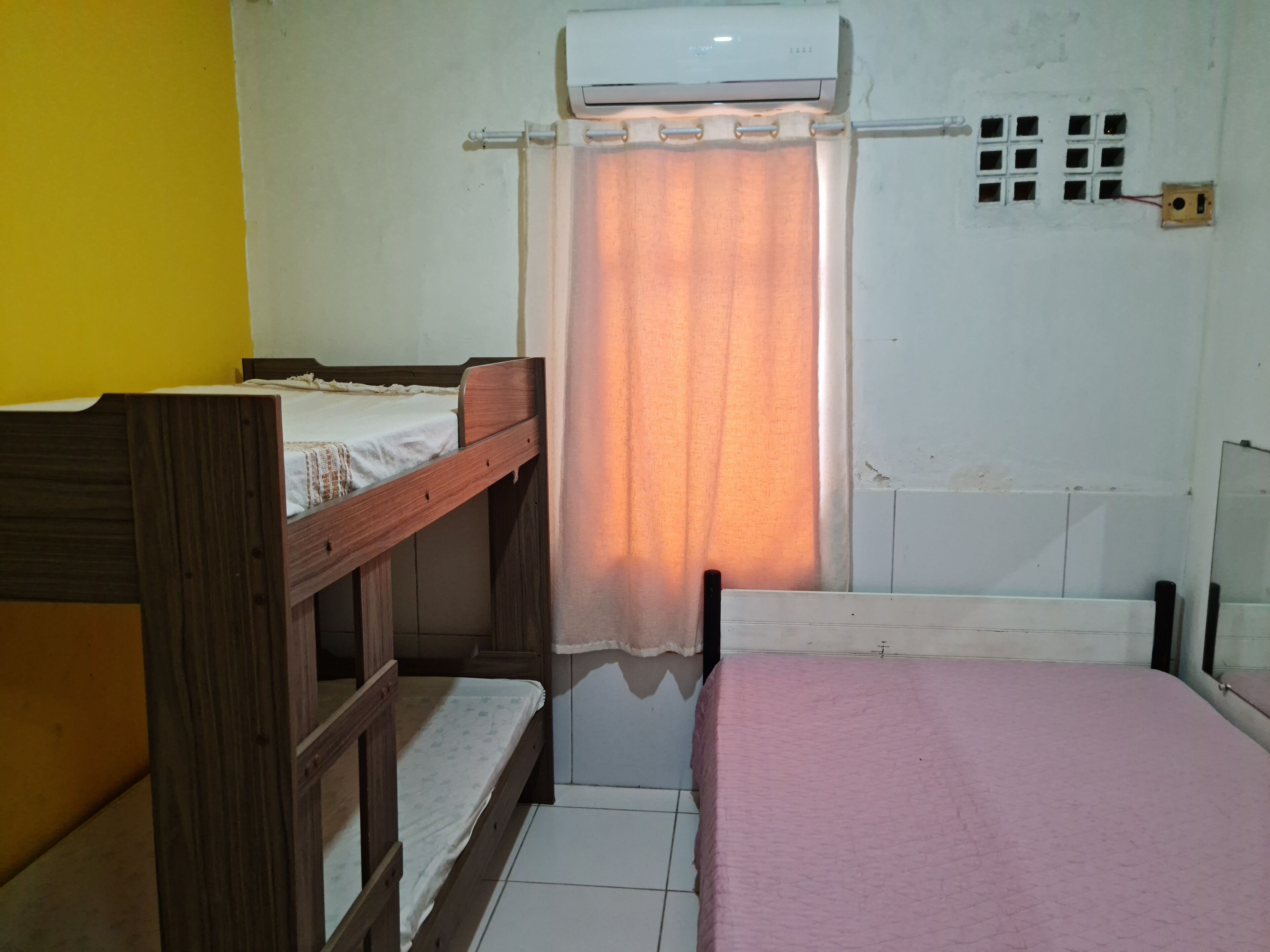 Une chambre de dortoir équipée de deux lits superposés, d’un lit simple, d’un appareil de climatisation, d’une fenêtre avec un rideau et d’une prise de courant murale.
