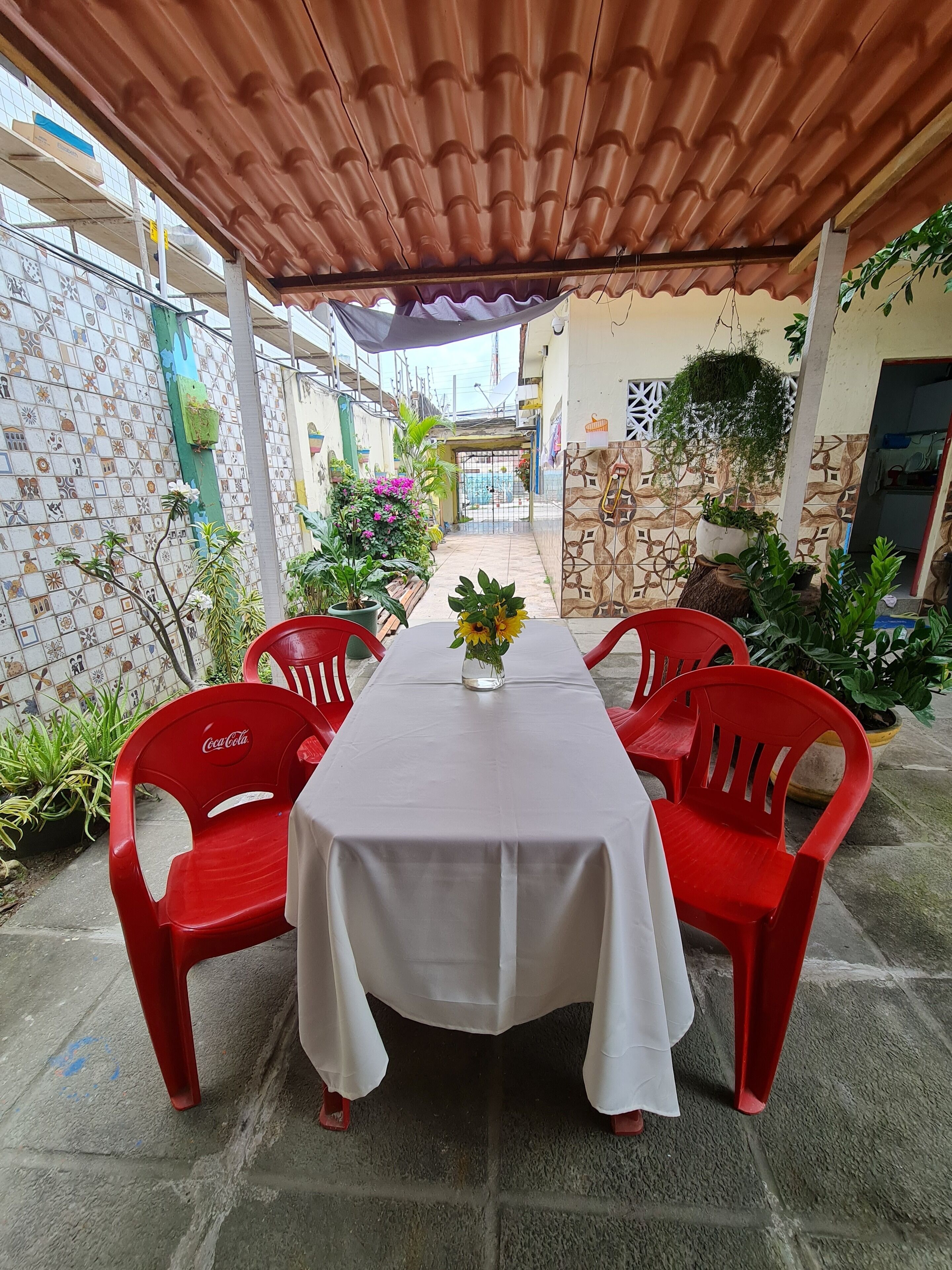 A covered patio with a table and red chairs, surrounded by potted plants and a tiled wall.