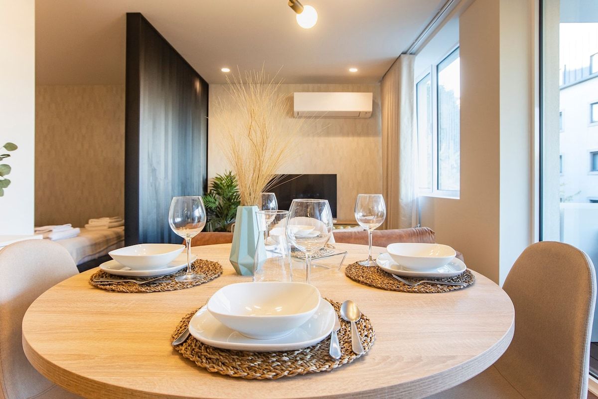 Ein runder Esstisch ist gedeckt mit weißen Tellern, Schüsseln und Weingläsern, mit modernem Interieur und Blick nach draußen.