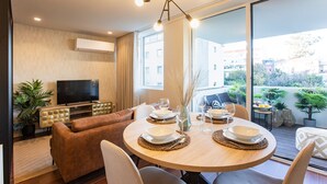 Ein modernes Wohnzimmer mit einem runden Esstisch für vier Personen, einem Ledersofa, einem Flachbildfernseher und einem Balkon mit Pflanzen.