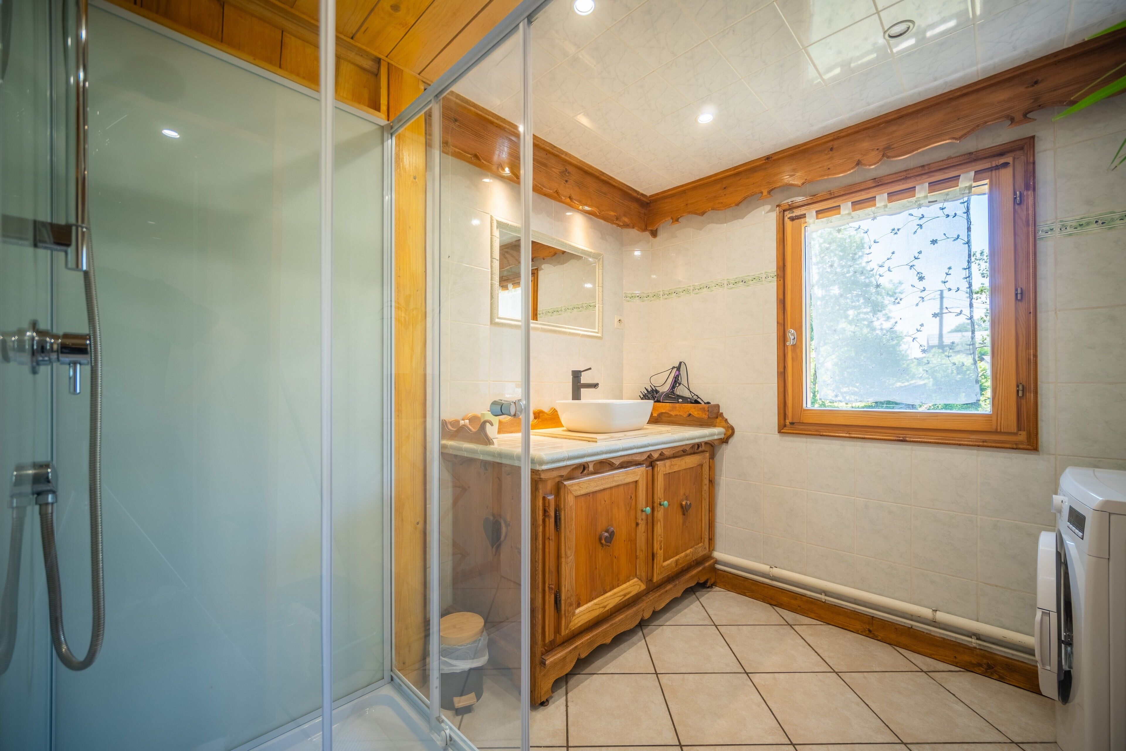 Une salle de bain dotée d’une douche à parois de verre, de meubles en bois, d’un lavabo avec un miroir et d’une fenêtre donnant sur un paysage verdoyant.