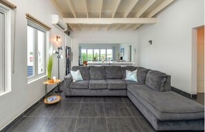 Ein modernes Wohnzimmer mit einem grauen Ecksofa, einem kleinen Beistelltisch und einem großen Fenster mit Jalousien.