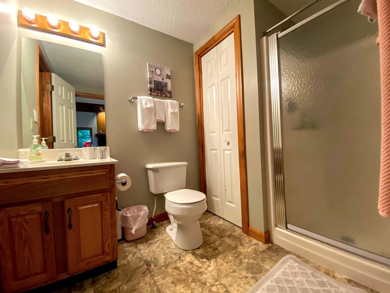 A bathroom with a wooden vanity, a toilet, a shower with a glass door, and a mirror with lights.