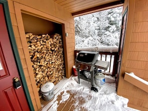 A wooden cabin entrance with a stack of firewood, a barbecue grill, and a snow-covered deck. - Ski Cabin Rental Ideal for a Mount Washington Vacation (Carroll)