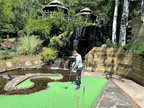 Iemand speelt minigolf in de buurt van een kleine waterval en gebouwen met rieten daken.