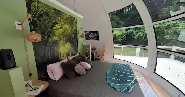 A room with a bed, a desk, a chair, a window with a view of trees, and a wall with a forest mural.