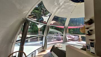 A room with a curved ceiling, a bed, a window, and a view of an outdoor area with a red umbrella.