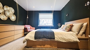 Un dormitorio con cama, cómoda, silla y ventana con vista a edificios.