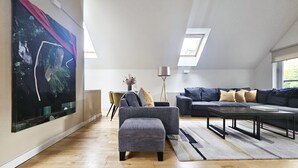 Un salón amplio con techo alto, un cuadro grande en la pared, un conjunto de sofás, una mesa de centro y un comedor con sillas y una lámpara.