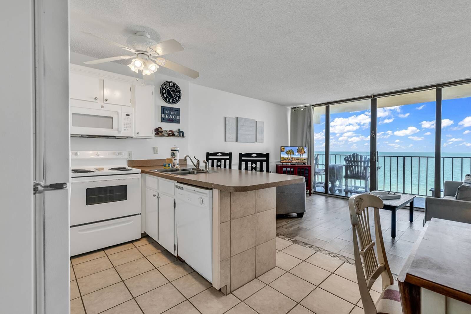 A kitchen with white appliances, a dining area with a view of the ocean, and a ceiling fan.