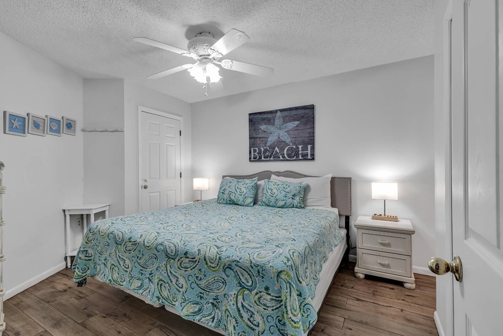 A bedroom with a bed, two bedside tables, a ceiling fan, and a wall with a 'BEACH' sign.
