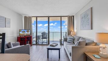 A spacious living room with a balcony overlooking the sea, a television, a red cabinet, and a comfortable seating area.