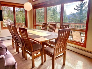 Un espace repas avec une table et des chaises en bois, de grandes fenêtres offrant une vue sur les arbres et les montagnes, et un luminaire suspendu.