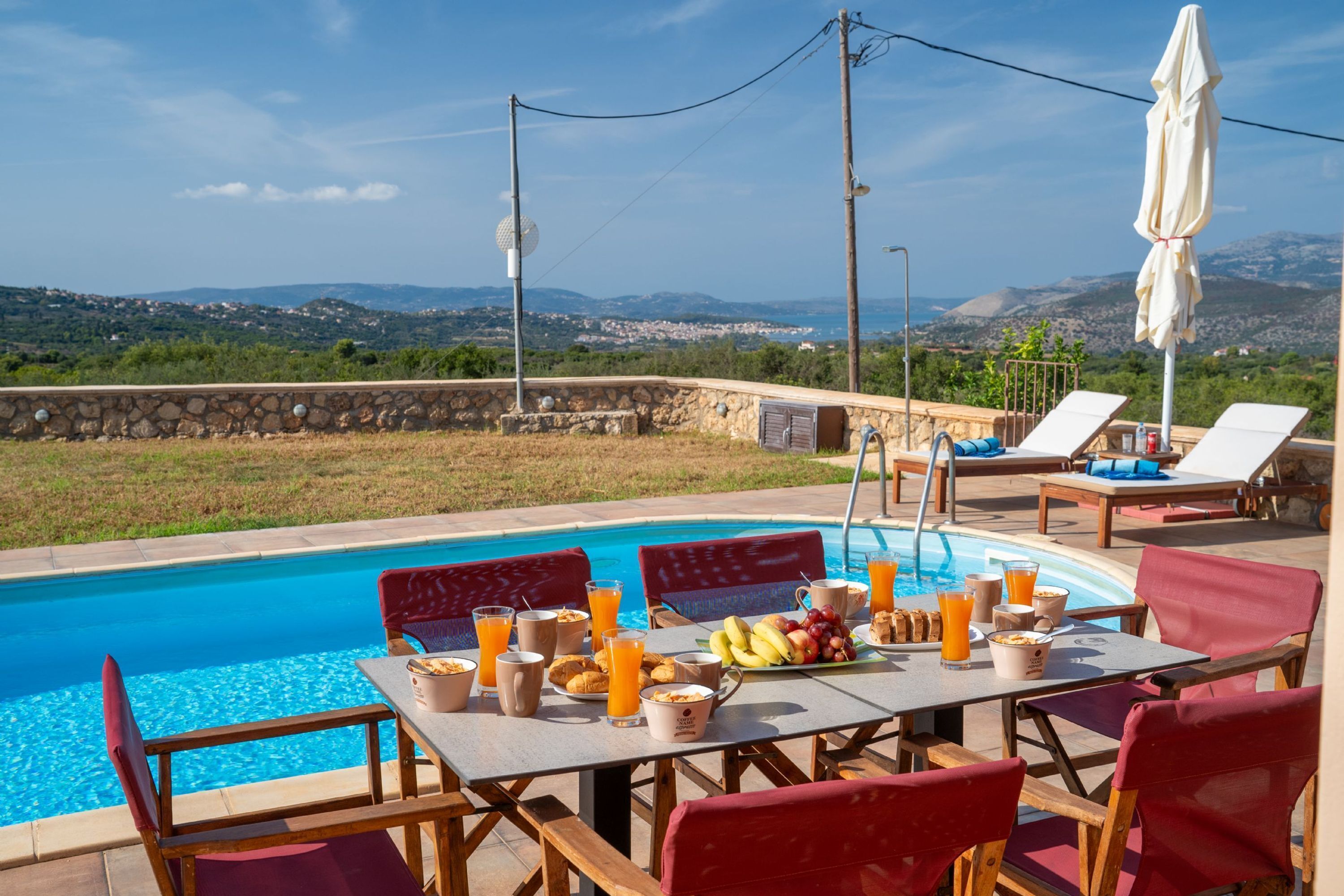 Área de refeições à beira da piscina, com mesa posta para o café da manhã, com vista para a piscina e colinas panorâmicas.