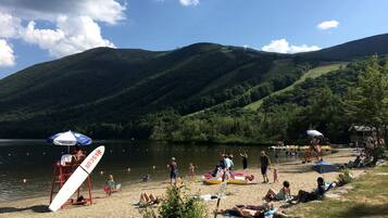 A lakeside beach with people relaxing, a lifeguard stand, and a mountain backdrop.