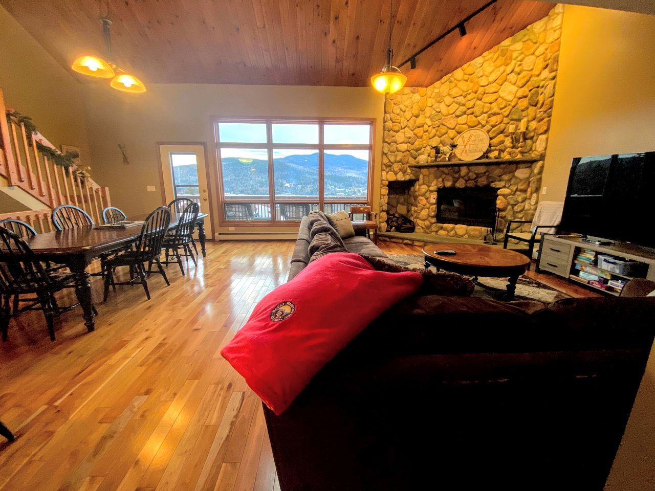 A cozy living room with a stone fireplace, wooden flooring, a dining area with chairs, and a large window offering a view.