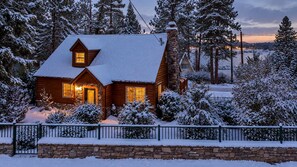 Une cabane en bois avec une cheminée en pierre, entourée d’arbres couverts de neige et d’un mur de pierres.