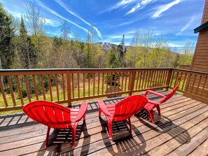 Eine Holzterrasse mit drei roten Stühlen und einem Tisch, mit Blick auf eine bewaldete Landschaft und einen Berg in der Ferne.