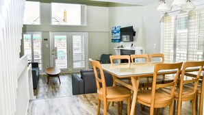 A dining area with a glass-top table and wooden chairs, a gray sofa, and a view of the outside through large windows.