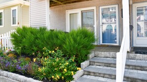 A two-story house with a front porch, a small garden with various flowers, and a set of stone steps leading to the front door.