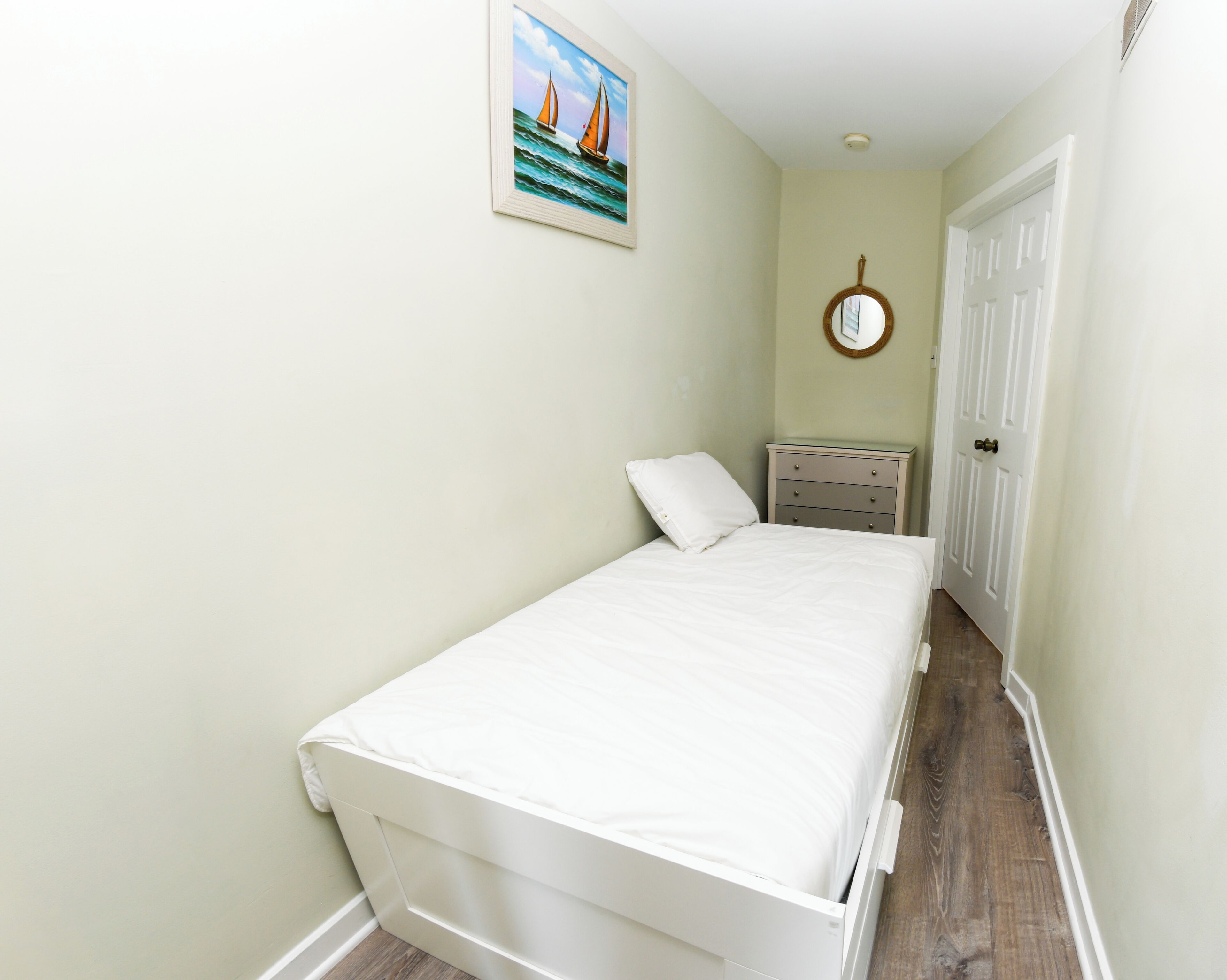 A narrow hallway leading to a bedroom with a bed, a dresser, a mirror, and a painting on the wall.