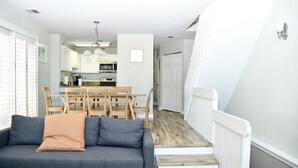 A modern living room with a grey sofa, wooden dining table, and chairs. A staircase leads to a second floor with a white railing.