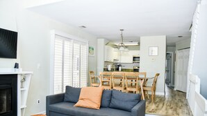 A living room with a gray sofa, wooden coffee table, and a fireplace. There is a dining area with a wooden table and chairs, and a kitchen in the background.
