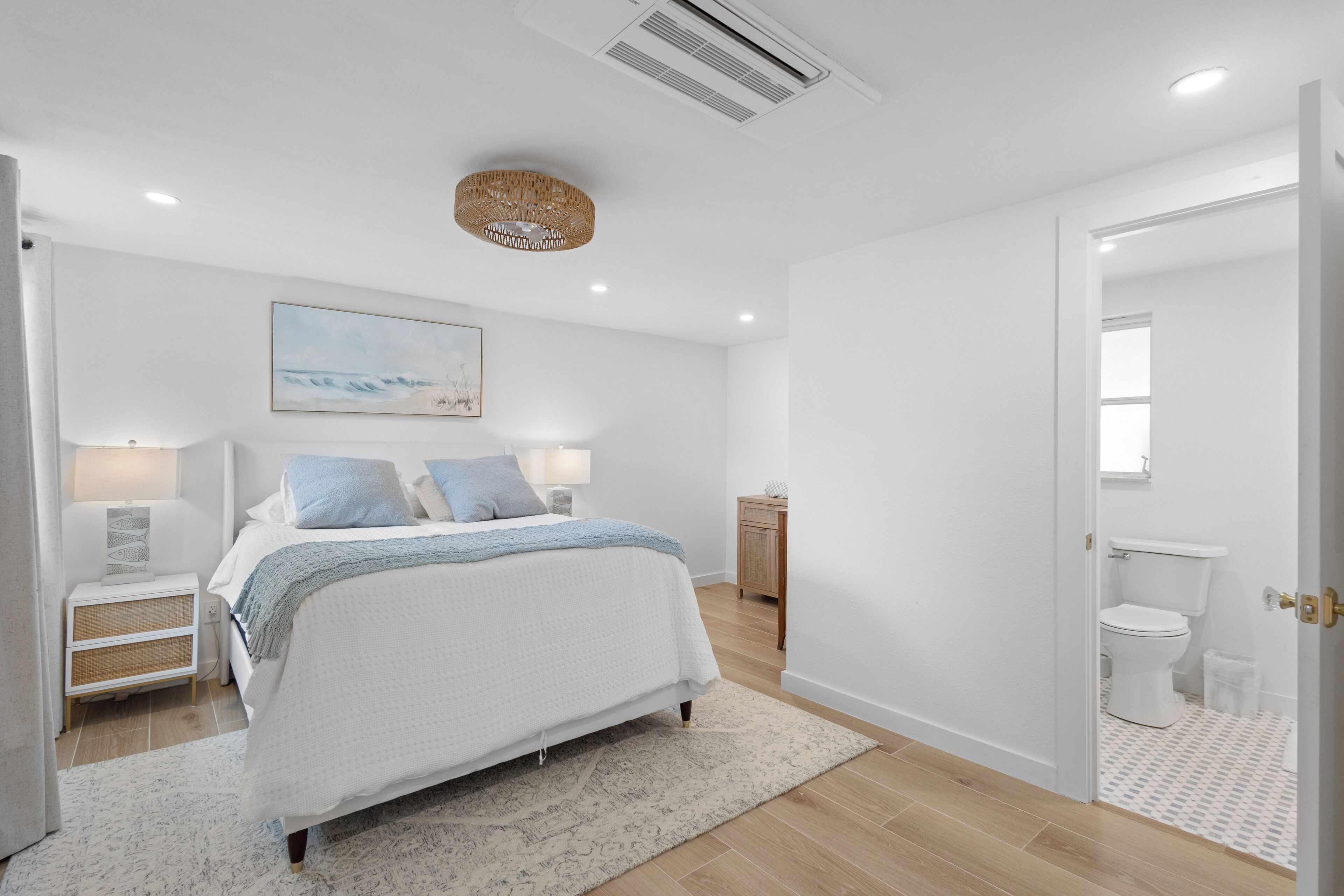 Ein Schlafzimmer mit einem Bett, Nachttischen, einem Wandkunstwerk, einem Deckenventilator und einer Tür zu einem Badezimmer.