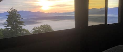 A room with a large window overlooking a mountainous landscape during sunset.
