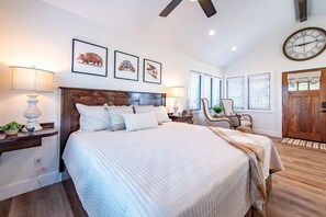 Une chambre à coucher avec un grand lit, une tête de lit en bois, des œuvres d’art encadrées accrochées au mur, une horloge et une porte en bois.