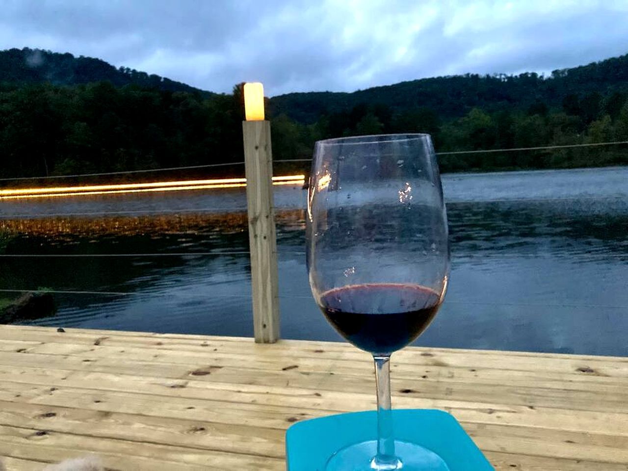 Ein Glas Rotwein auf einer Holzterrasse mit Blick auf einen See und Hügel.