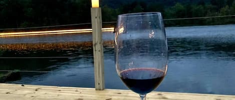 A glass of red wine on a wooden deck with a view of a lake and hills.