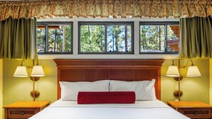 A hotel room with a large bed, two bedside tables, two lamps, and a view of trees through three windows. - Juniper-Boardwalk by Roundhouse Resort and Spa (Pinetop Country Club)