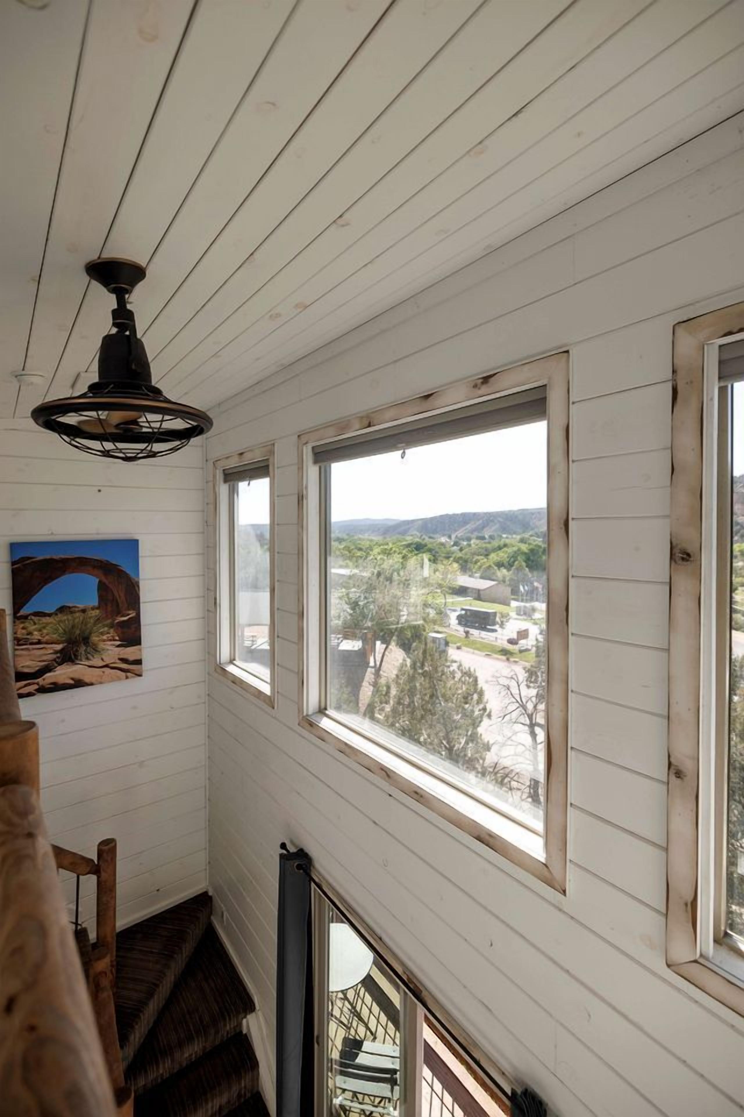 A room with white wooden walls, a ceiling fan, a staircase, and large windows offering a view of a landscape with trees and buildings.
