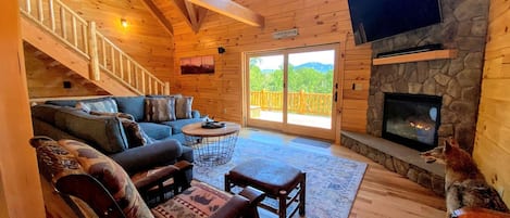 A log cabin interior with a stone fireplace, a television mounted on the wall, a staircase leading to an upper level, and a large sliding glass door to the outside.