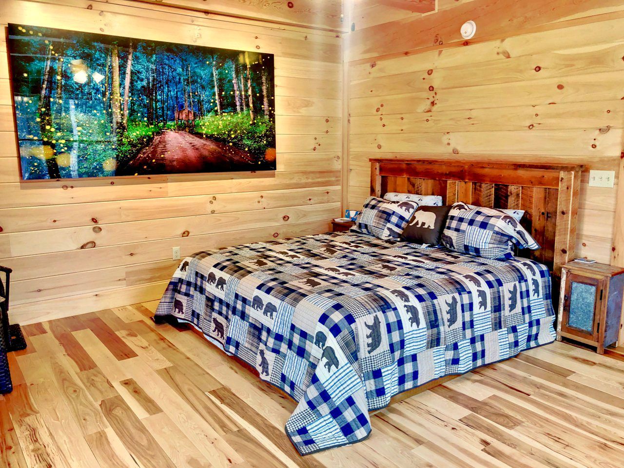 A wooden cabin bedroom with a bed, a painting of a forest, and a small cabinet.