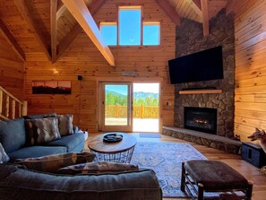 Interior de una cabaña de troncos con chimenea de piedra, un sofá, una mesa de centro y un televisor grande montado en la pared.