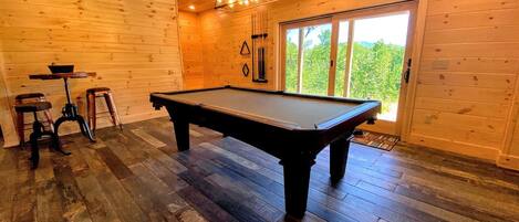 Una mesa de billar en una habitación con paredes y suelos de madera, una lámpara en el techo y una puerta que da al exterior.