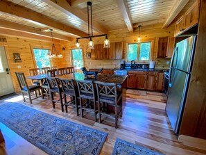 A wooden cabin interior with a dining area, kitchen, and large windows.