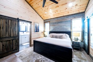 A bedroom with a large bed, wooden walls, a ceiling fan, and a view of the outside. - Contemporary Coziness Cabin in Hochatown, OK (Hochatown)