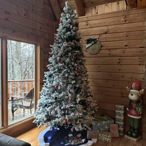 Ein Holzblockhaus-Innenraum mit einem geschmückten Weihnachtsbaum, einem Fenster mit Blick auf Bäume und einem Kuscheltier auf dem Boden.