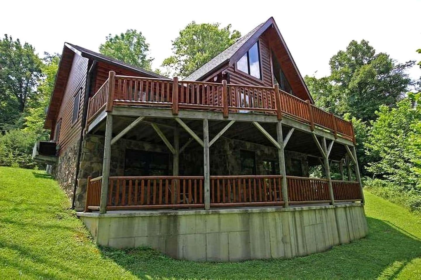 A wooden cabin with a balcony, stone foundation, and surrounded by greenery.
