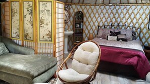 A traditional yurt interior with a bed, a cushioned chair, a small shelf, and a decorative screen. - Rustic Luxury: Riverfront Yurt Glamping in Claxton, Georgia (Claxton)