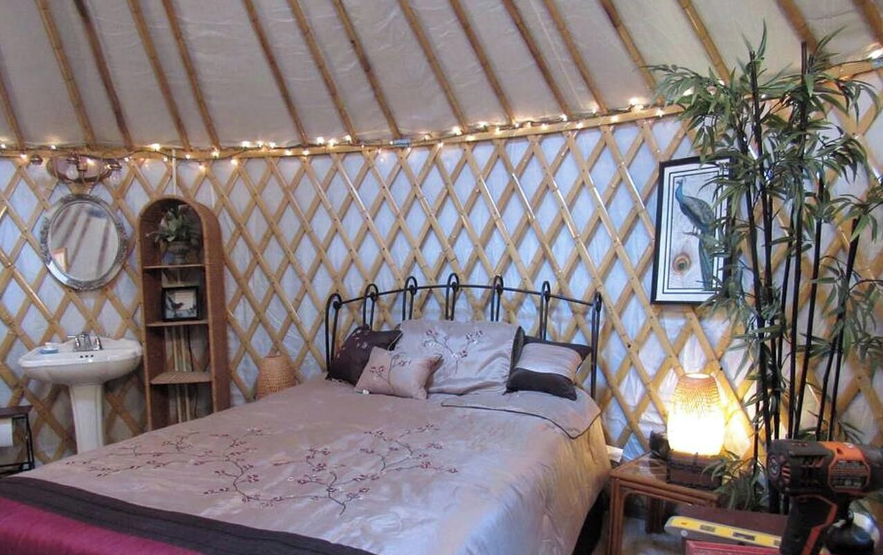 A bed with a headboard, a bedside table, a lamp, a mirror, a shelf, and a potted plant in a tent-like room.
