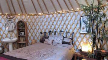 A bed with a headboard, a bedside table, a lamp, a mirror, a shelf, and a potted plant in a tent-like room.