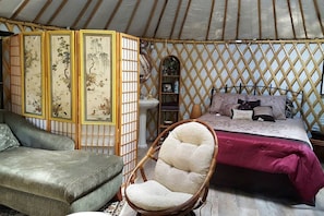 Ein traditionelles Jurten-Interieur mit einem Bett, einem gepolsterten Sessel, einem kleinen Regal und einem dekorativen Raumteiler.