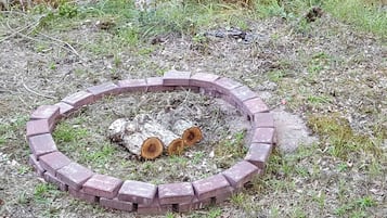 Un foyer circulaire en briques, avec des bûches au centre, entouré d’herbe et de mauvaises herbes.