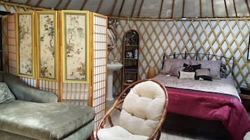 L’intérieur d’une yourte traditionnelle est aménagé avec un lit, un fauteuil rembourré, une petite étagère et un paravent décoratif.